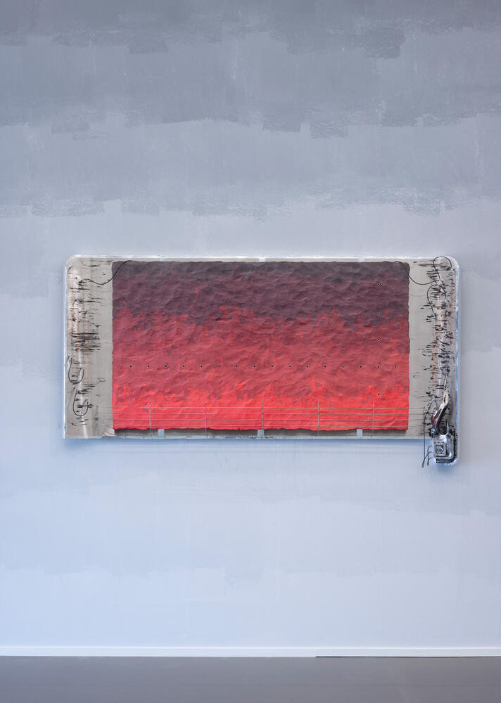 An der Wand h&auml;ngt ein Kunstwerk. Die Seiten sind grau, aus denen allerlei elektrische Dr&auml;hte ragen. Die mittlere Fl&auml;che besteht aus verschiedenen Rott&ouml;nen.