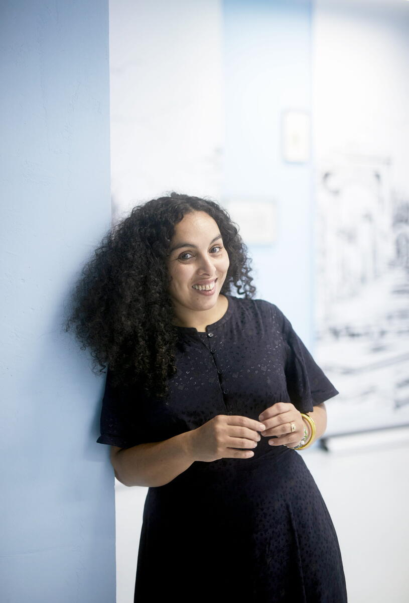 Chourouk Hriech leans against a pale blue pole. She wears a black dress and has black curly hair. She smiles.