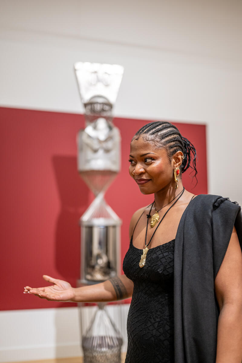 Kunstenaar Minne Atairu staat met haar handpalm open. Ze glimlacht en kijkt naar de verte. Achter haar is een ronde achtergrond en metalen kunstwerk.