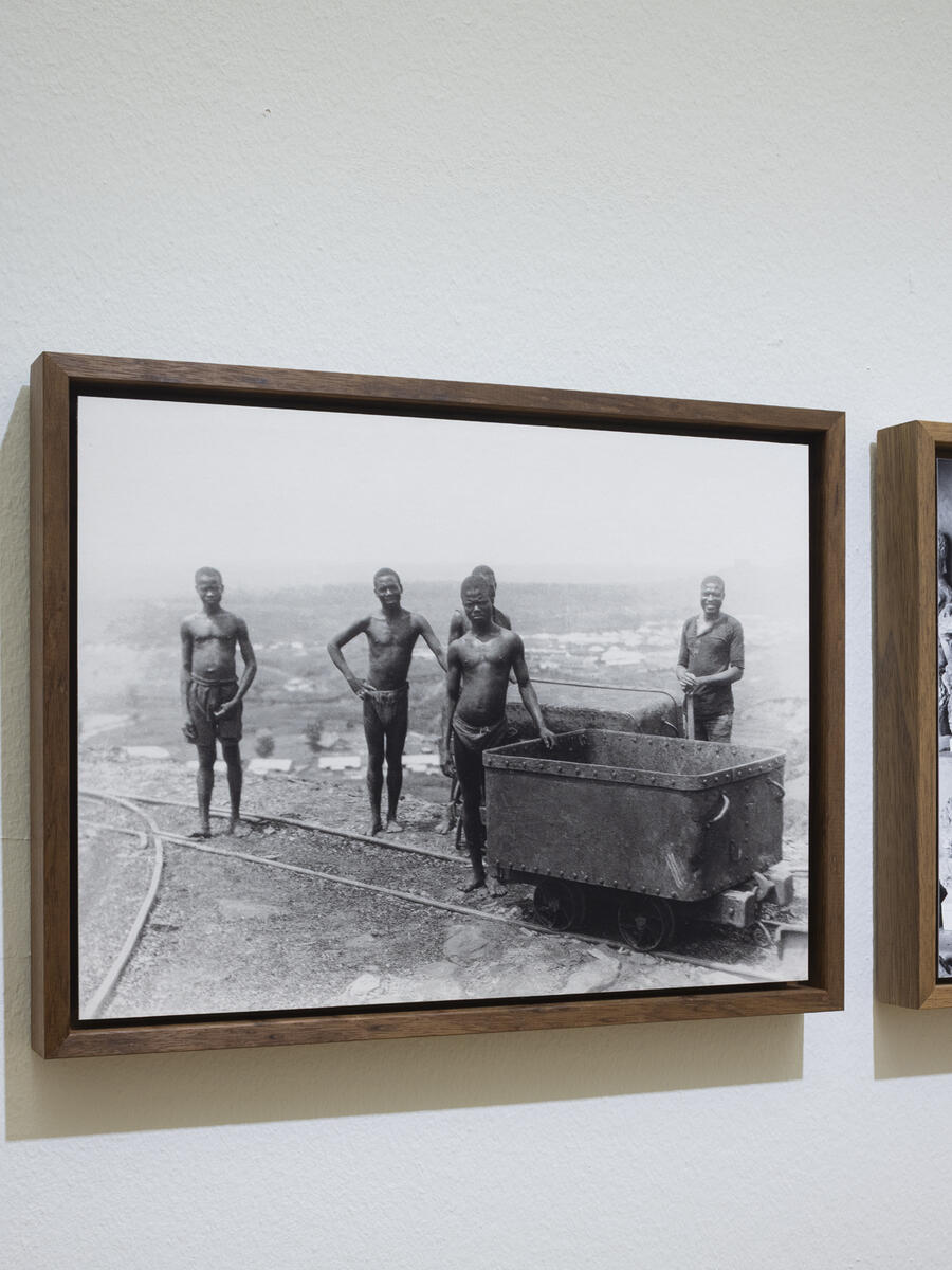 Zwart-wit foto van arbeiders, vermoedelijk in Nigeria. De jonge mannen staan halfnaakt op de foto naast een kar op rails, vermoedelijk voor het vervoeren van brons.