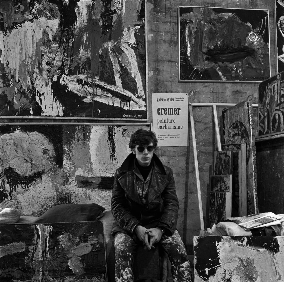 A black-and-white photograph of a young Jan Cremer. He wears sunglasses and sits on a stool, surrounded by paint stuff.