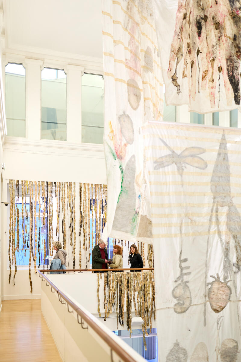 Menschen stehen auf der Balustrade und unterhalten sich miteinander. Von der Decke h&auml;ngen textile Kunstwerke in Wei&szlig; und Blassrosa.