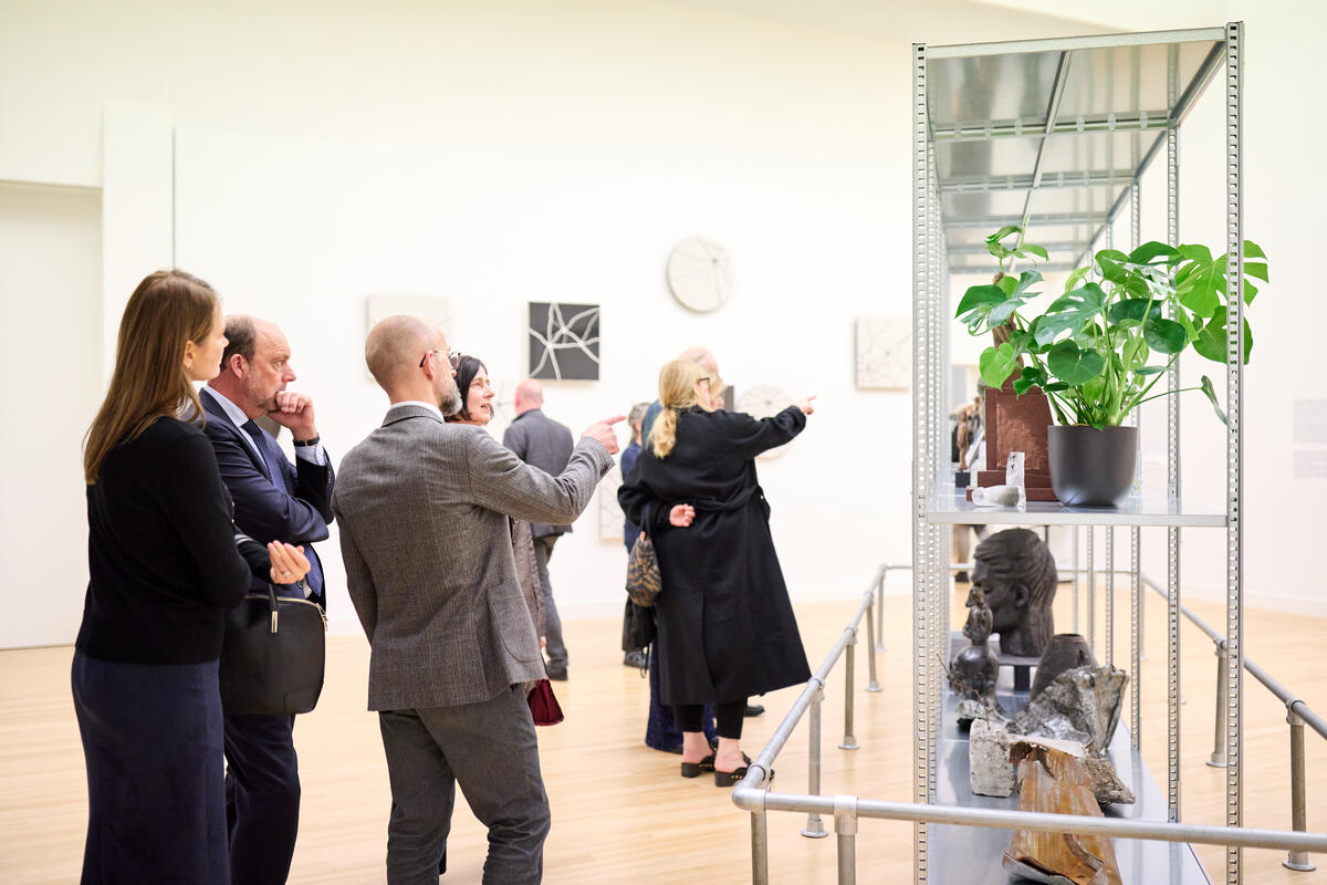 Menschen in Anz&uuml;gen stehen vor einem Kunstwerk und unterhalten sich miteinander.