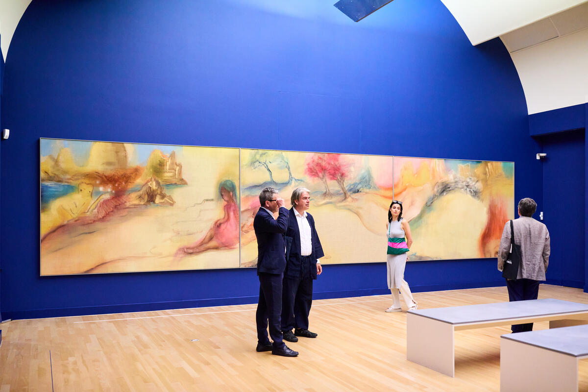 Verschillende bezoekers, waaronder mensen in pak, staan in het museum. De muur achter hen is blauw. Op de muur hangt een groot schilderij in pastelkleuren.