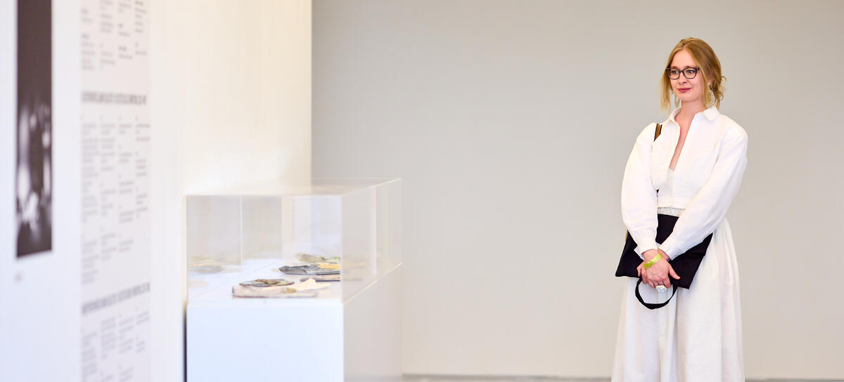 Een vrouwelijke bezoeker in witte jurk staat bij een witte muur met informatie over een expositie. Er staat een witte vitrinekast met kunstwerken erin die niet goed zichtbaar zijn. Alles in de foto is in wit tinten.