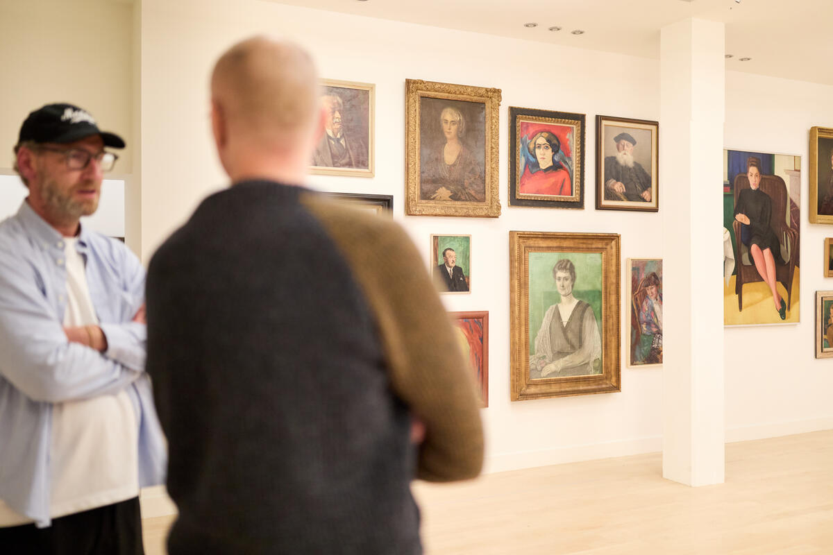 Zwei m&auml;nnliche Besucher unterhalten sich in einem Raum, in dem Portr&auml;ts von Frauen an der Wand h&auml;ngen. Die M&auml;nner sind unscharf.
