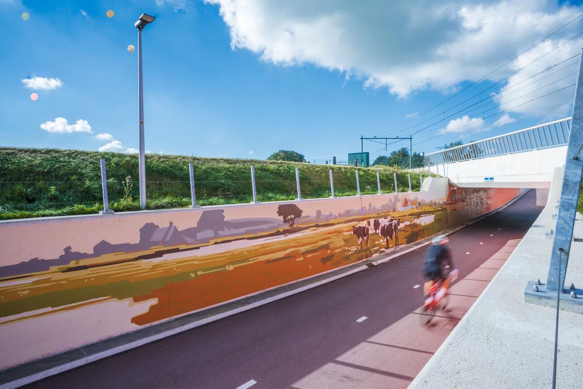 Een fietser fietst door een fietstunnel met daarop een muurschilderij van een landschap in zachte kleuren. De lucht is blauw.