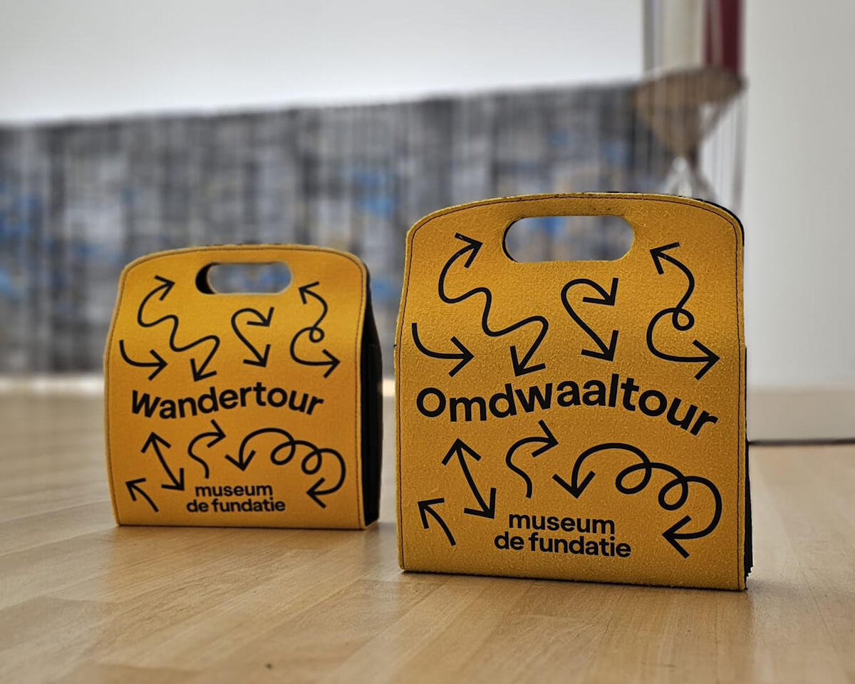 Twee gele tassen staan in het museum. Op de tassen staat omdwaaltour en wandertour. Er staan allemaal grappige pijltjes op de tassen.