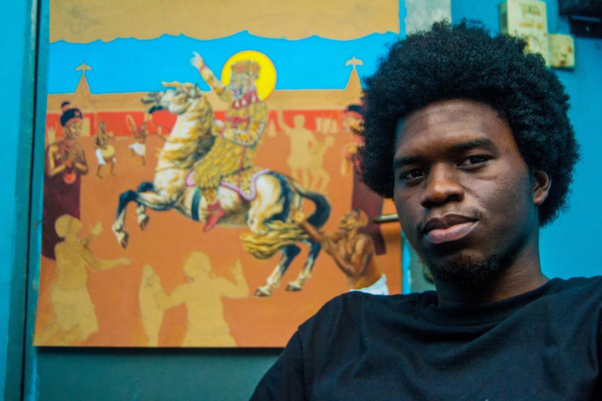 Portretfoto van Osaze Amadasun. Hij staat naast een schilderij in blauw en oranje van een ridder op een paard.