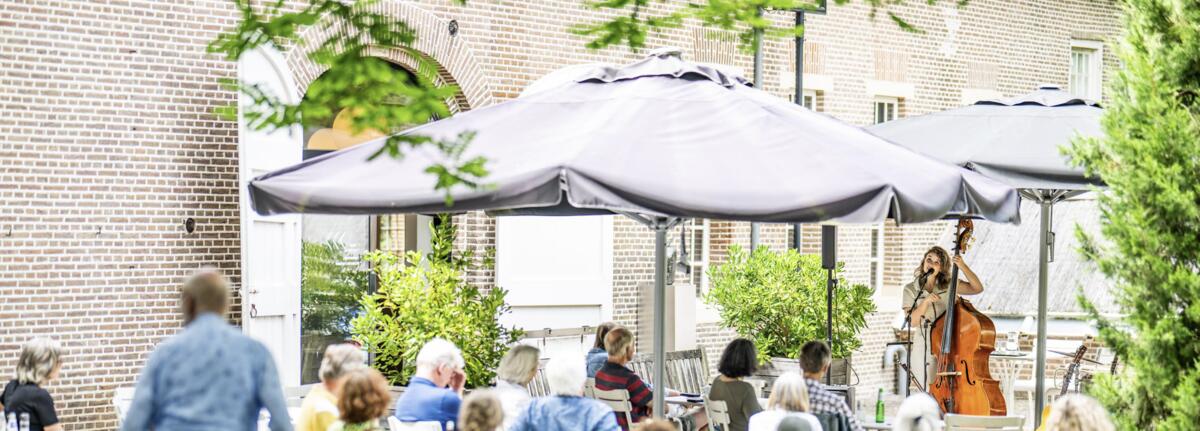 Die Menschen sitzen auf einer Terrasse und h&ouml;ren einer Frau zu, die Kontrabass spielt und singt.
