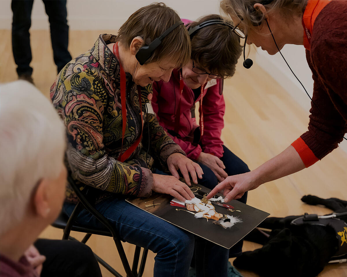 Twee mensen met een visuele beperking gaan met hun handen over een voel-schilderij. Een museumdocent staat erbij en legt wat uit.