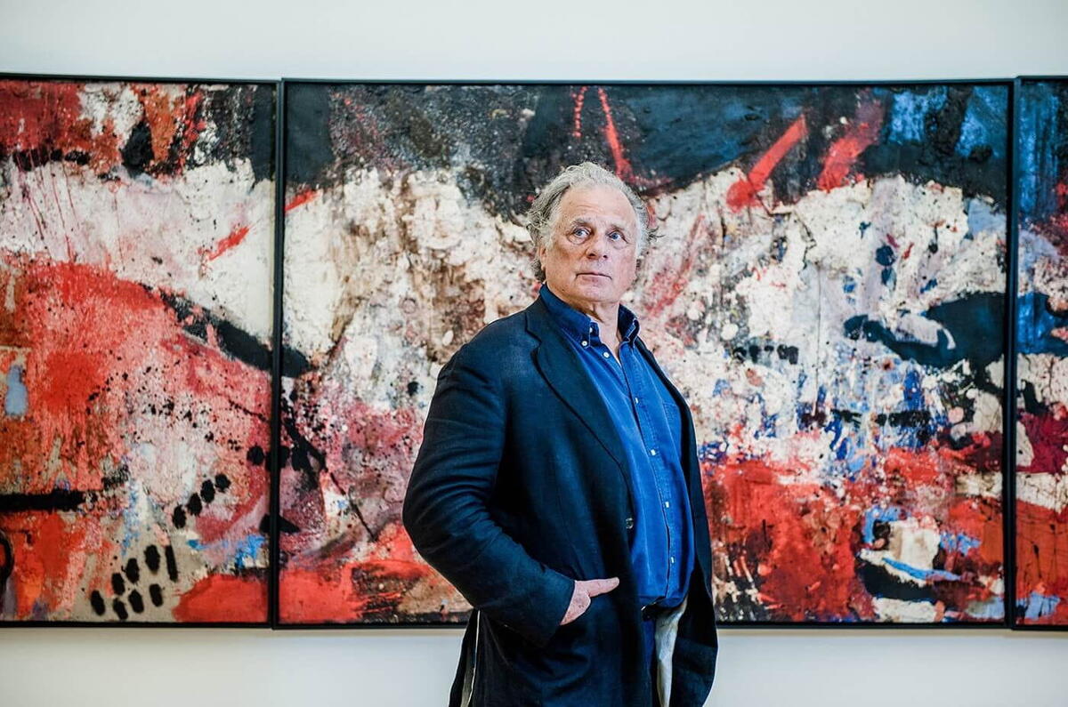 An elderly Jan Cremer stands in front of a large painting in red and dark blue. Cremer himself is wearing a dark blue suit and looking into the distance.