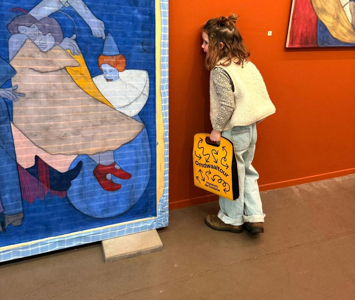Een jong meisje staat met haar wang tegen de muur en kijkt naar de achterkant van een schilderij. In haar hand draagt ze een gele tas waar Omdwaaltour opstaat. 
