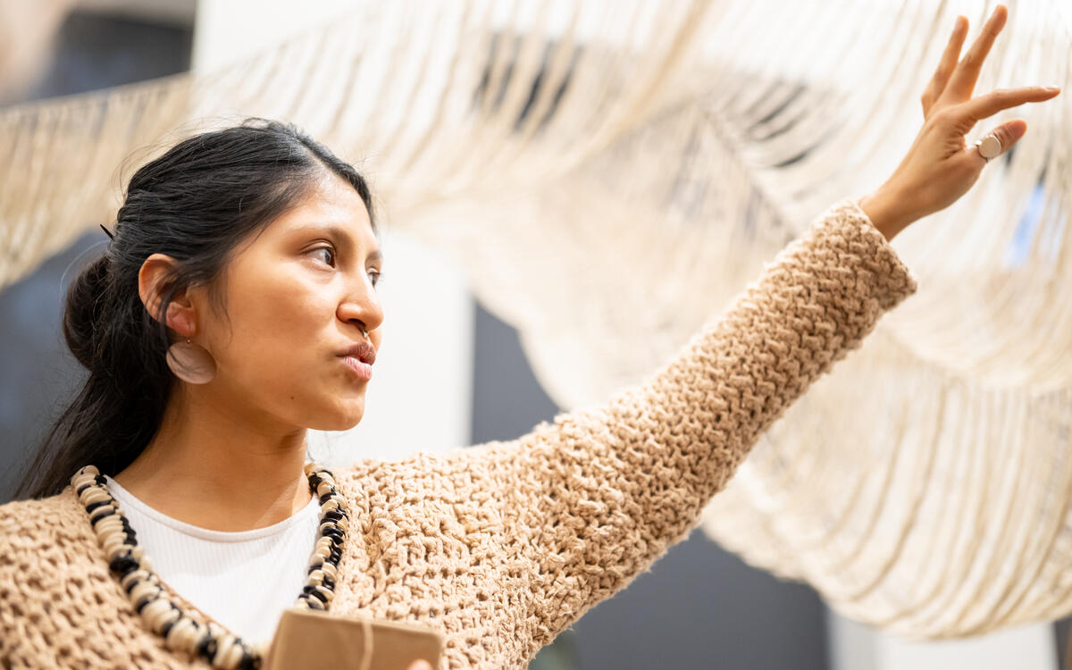 een close-up foto van Cristina Flores Pescorán, ze houdt haar arm omhoog. In de achtergrond zie je haar zandgekleurde geweven werk
