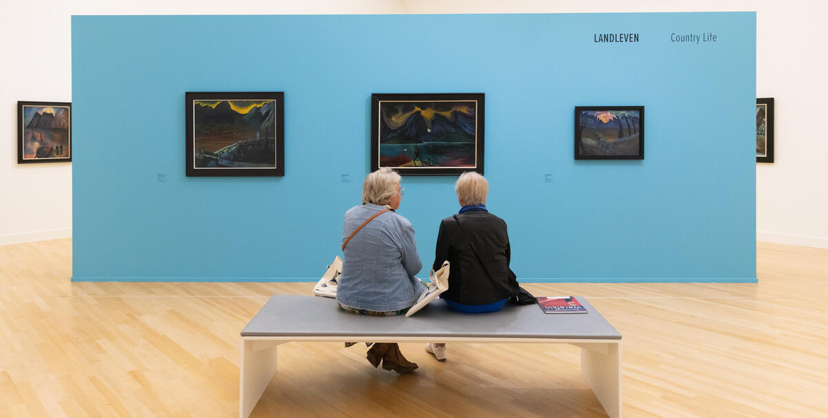 Twee mensen die met hun rug naar ons toe op een bankje zitten. Ze kijken naar een losstaande muur met daarop drie kunstwerken uit de Reset tentoonstelling.