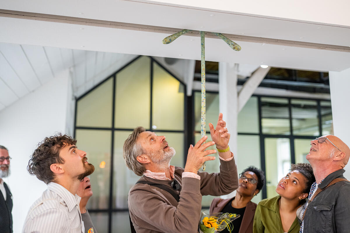 Een man wijst naar een kunstwerk dat van het lage plafond hangt. Een groep mensen kijkt mee omhoog, terwijl de man dingen uitlegt.