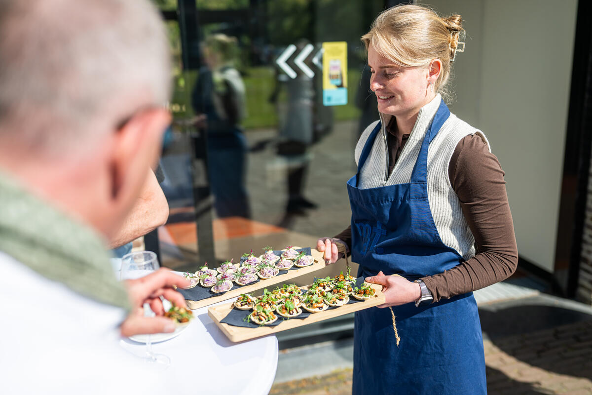 Een vrouw staat met een dienblad vol eten te glimlachen. Ze draagt een schort.
