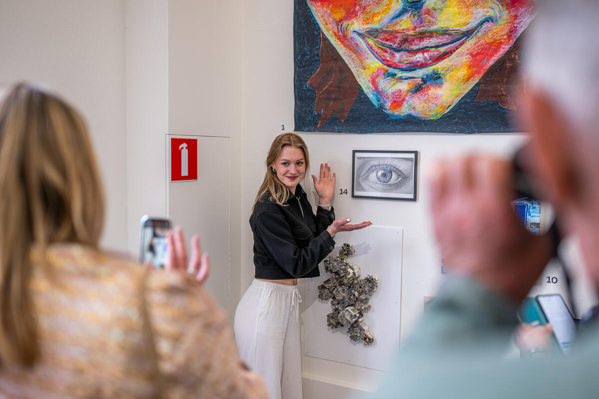 Een scholier staat bij een klein kunstwerk en lacht terwijl ze op de foto wordt gezet door anderen, terwijl ze naar het schilderij wijst.