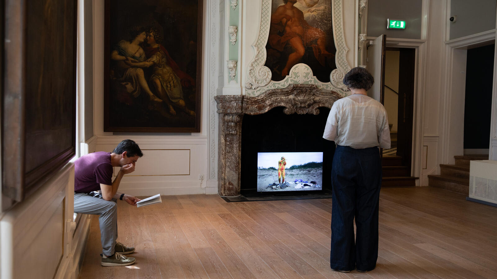 Een bezoeker zit in de vensterbank van een statige ruimte. Hij leest in een boekje. Op alle muren hangen kunst. Een vrouwelijke bezoeker staat en kijkt naar een beeldscherm waar een film op afspeelt.