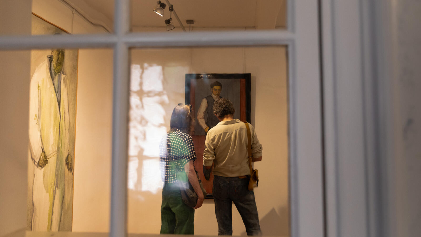Twee bezoekers staan voor een schilderij van een man. Links hangt nog een groot schilderij van een staande man. De foto is genomen door een raam.