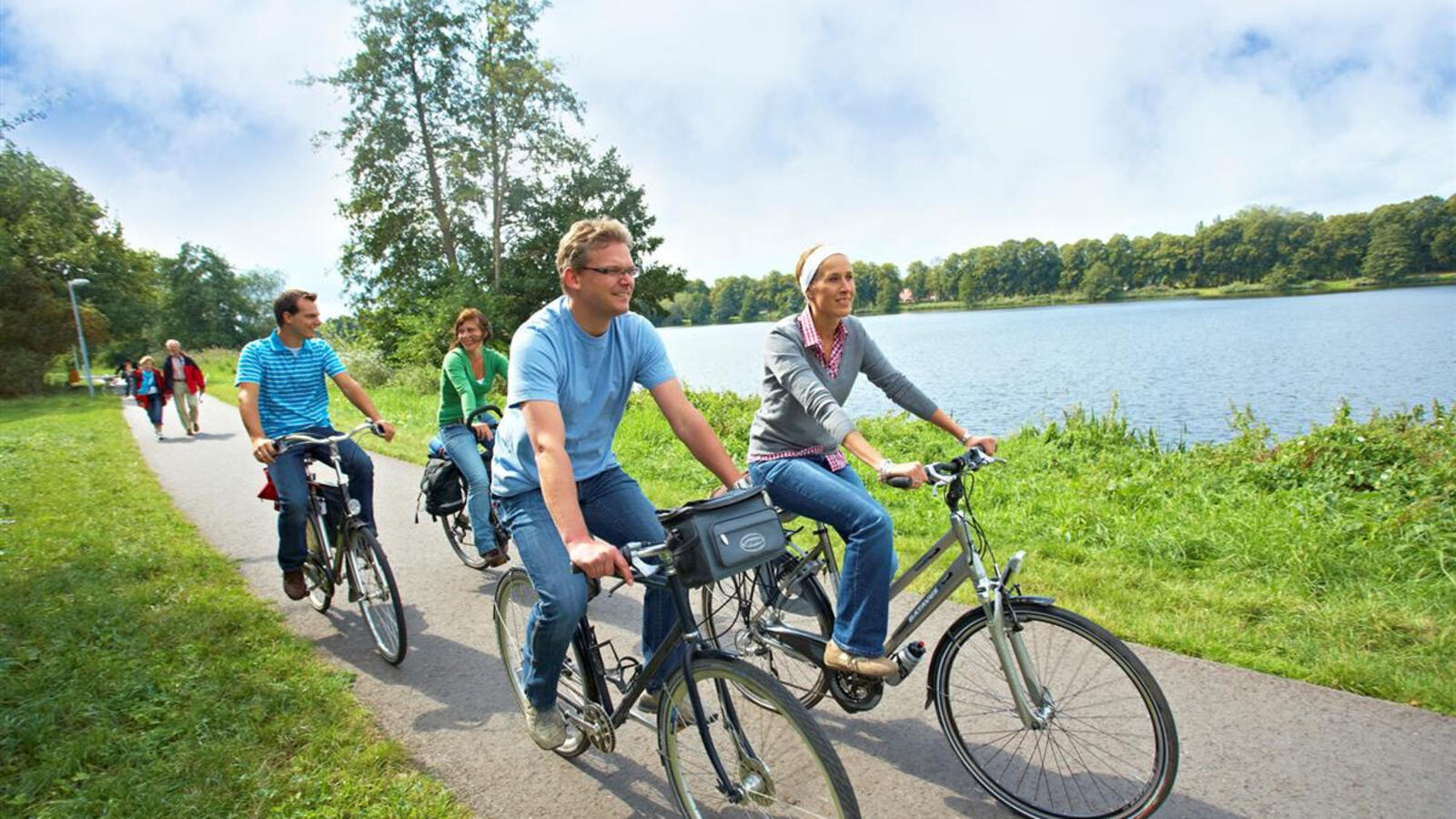 Mensen zijn aan het fietsen langs de Vecht, als onderdeel van de kunstroute. Ze lachen naar elkaar en zijn in gesprek. Achter de fietsers, in de verte, zijn een groepje mensen aan het wandelen.