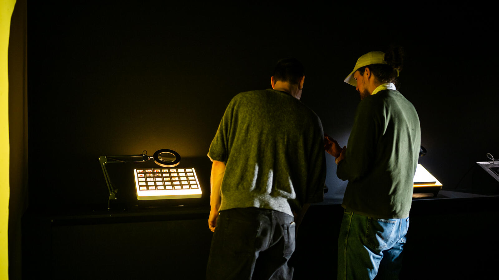 Twee jonge mannen staan met hun rug over een digitaal kunstwerk gebogen dat licht geeft. De ruimte is verder donker.