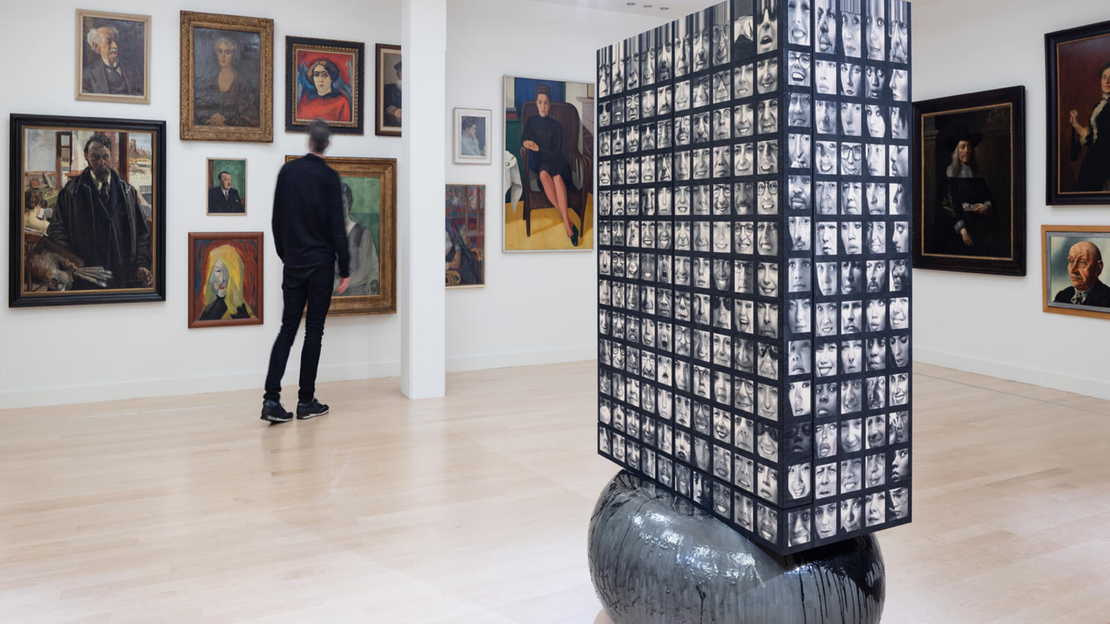 Een pilaar vol kleine foto's staat in een museumzaal. De muren achter de pilaar hangen vol met schilderijen.