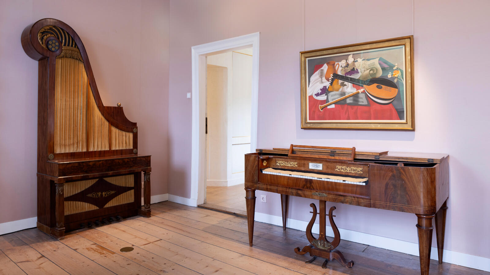Een antieke kast in de vorm van een rechtstaande pianovleugel en een oud bureau staan in een lichtroze kamer. Boven het bureau hangt een schilderij.