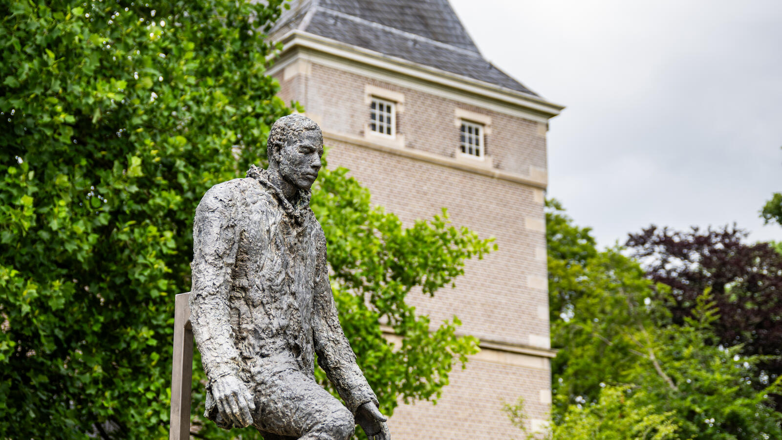 Een standbeeld van een man staat op de voorgront. Achter het standbeeld staat een grote boom en is een toren van Kasteel het Nijenhuis zichtbaar.