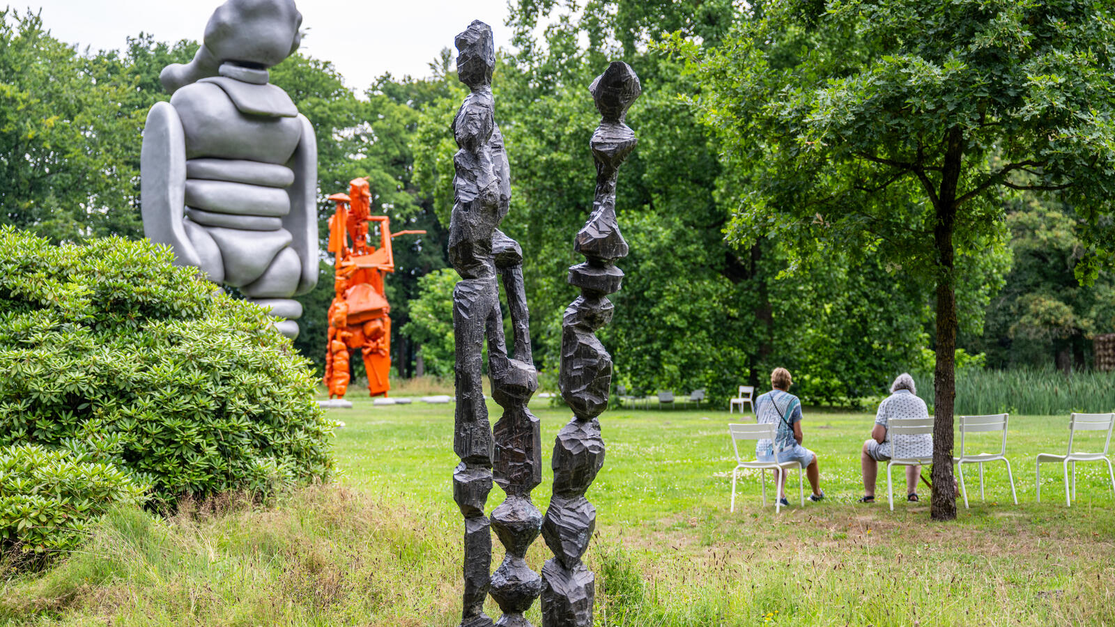 Een vergezicht van de beeldentuin. Er staan drie sculpturen, een dichtbij en twee in de verte. Onder een boompje zitten twee bezoekers op een bankje.
