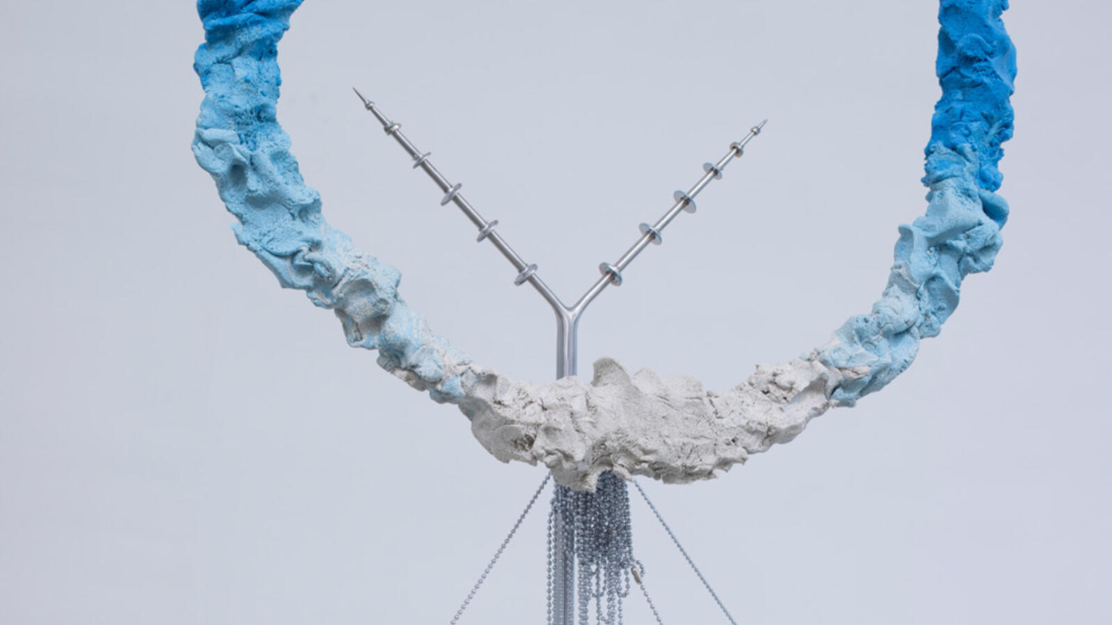 Een sculptuur met een grote ruwe cirkelvorm in wit en blauw, bevestigd op een witte sokkel met dunne metalen stangen, kettingen en puntige metalen elementen.