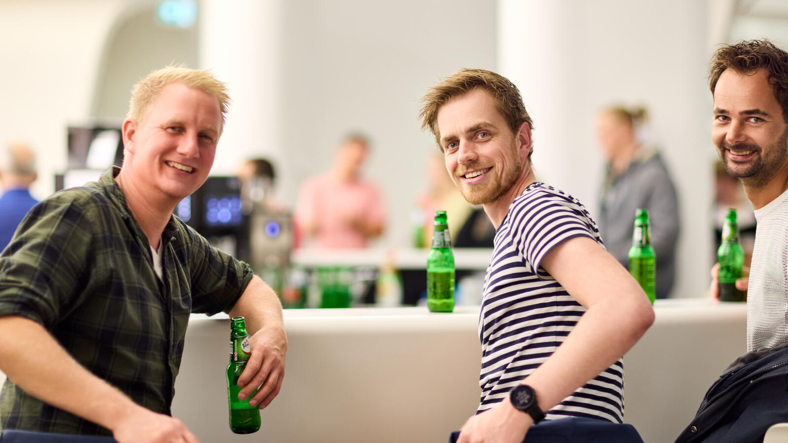 Drei junge Besucher sitzen mit einem Bier an der Bar des Museums. Sie drehen sich in die Kamera und lachen.