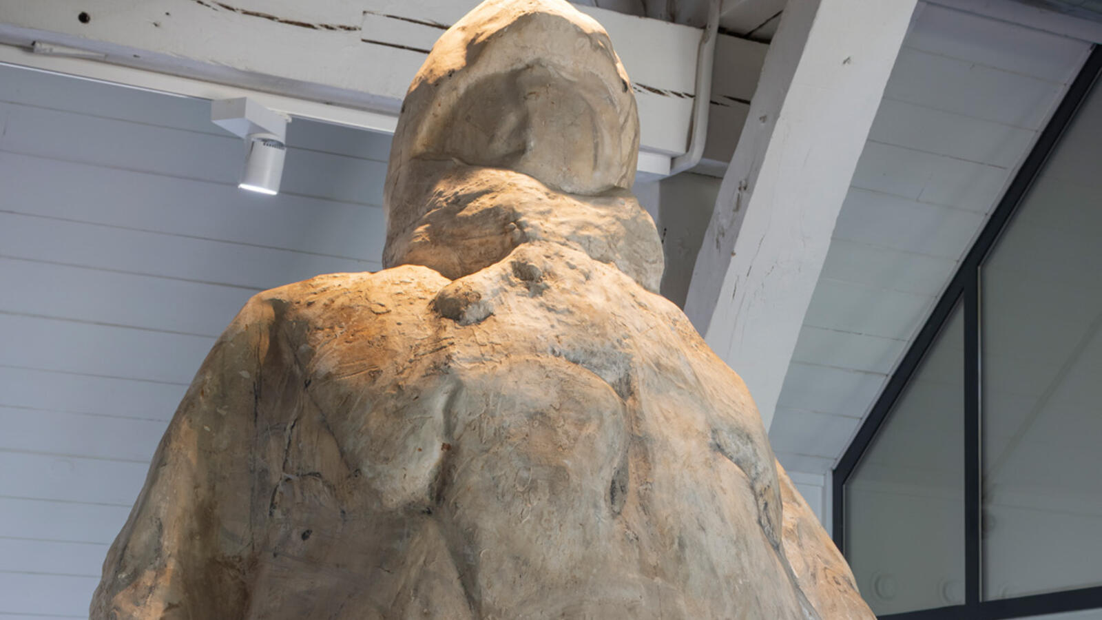 Een groot beeldwerk van steen van een rond mensachtig figuur staat onder een roestige lamp.