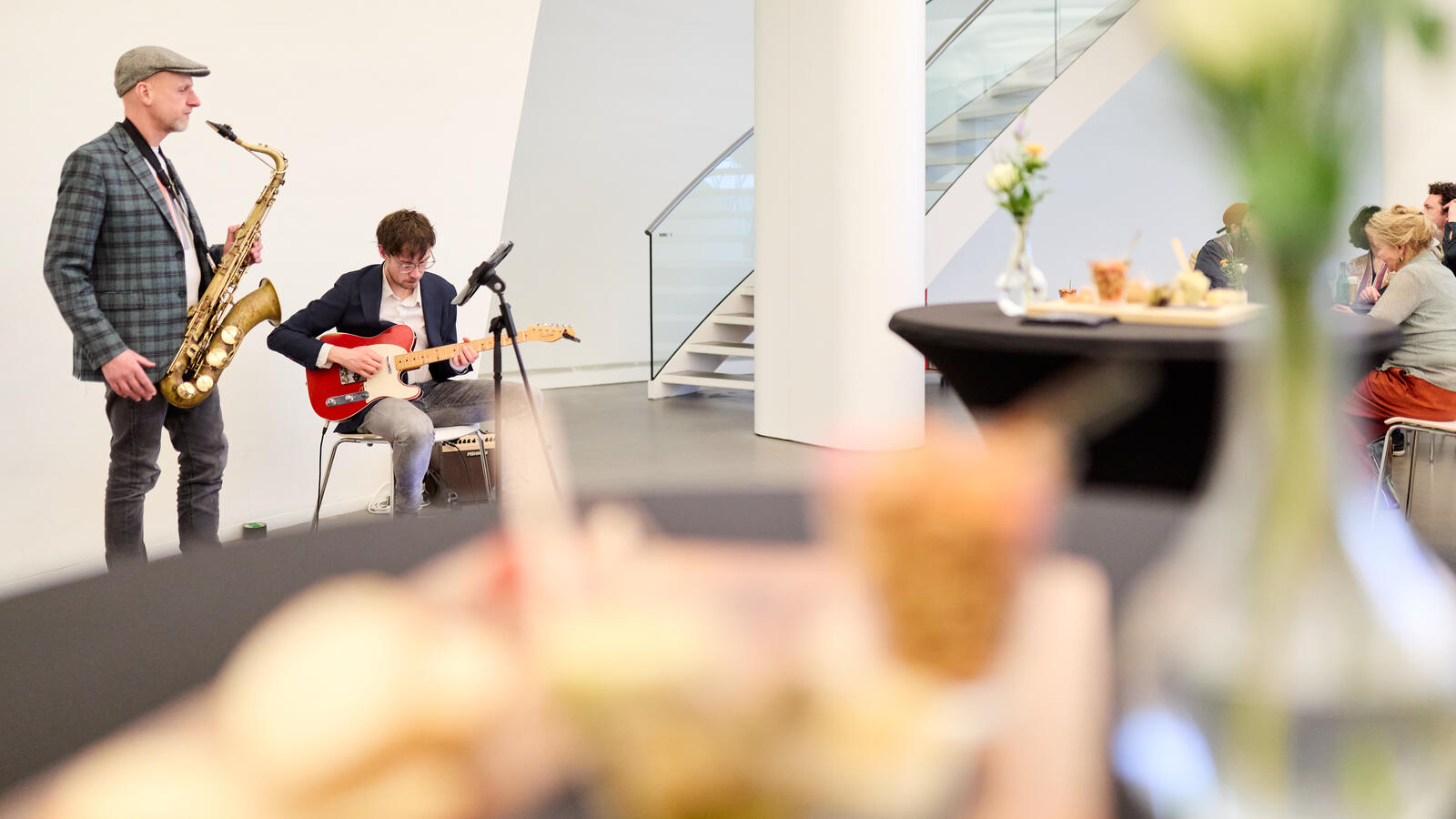 Een saxofonist en guitarist spelen in de Fundatie, terwijl mensen luisteren.