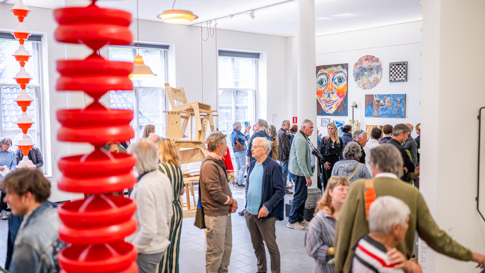 Tientallen bezoekers staan in de expositieruimte. Aan de voorkant van de foto hangt een rood kunstwerk van de plafond. 