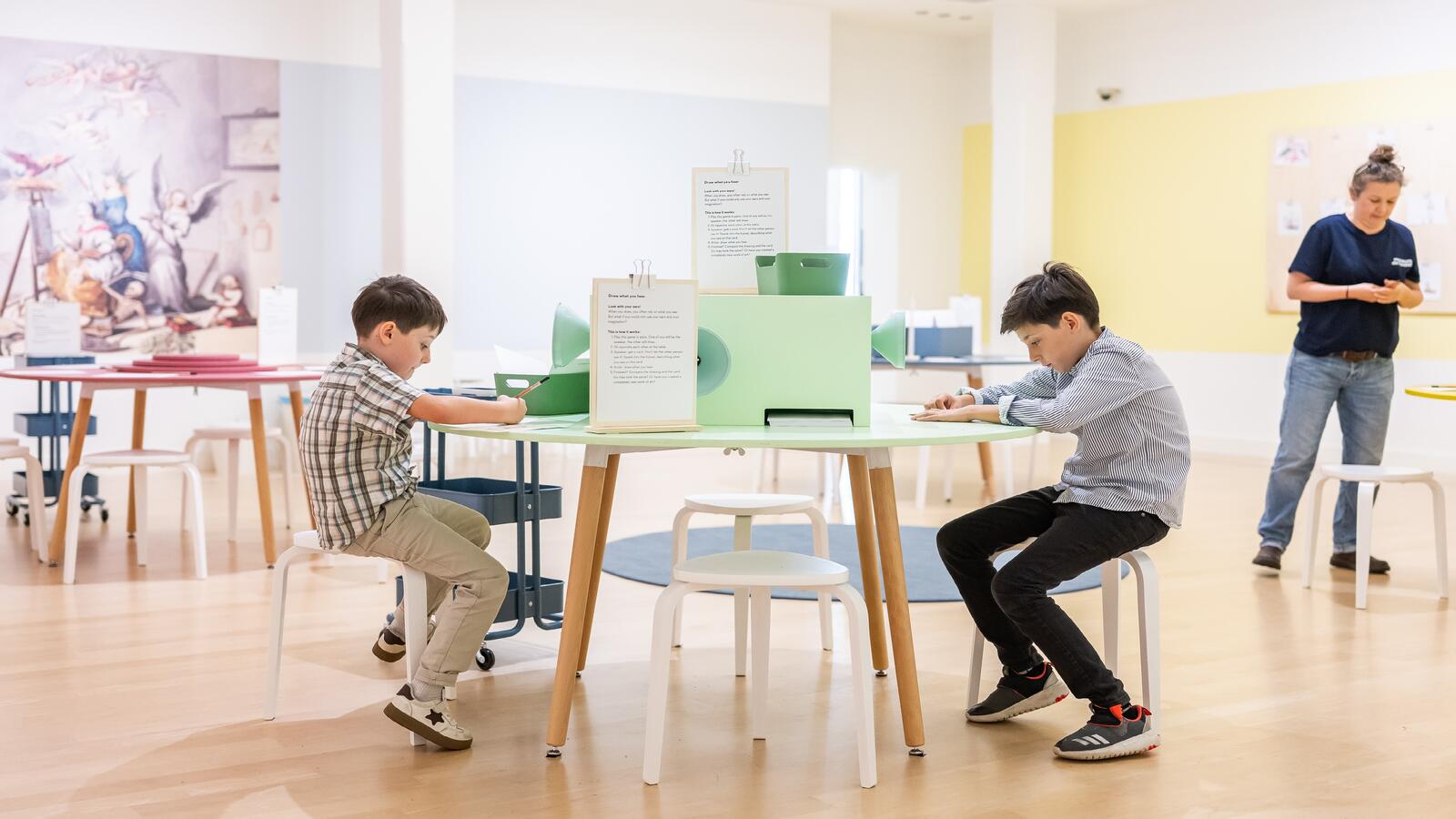 Twee jongens zitten in De Studio aan een ronde tafel te tekenen. Een vrouw staat bij een andere tafel.