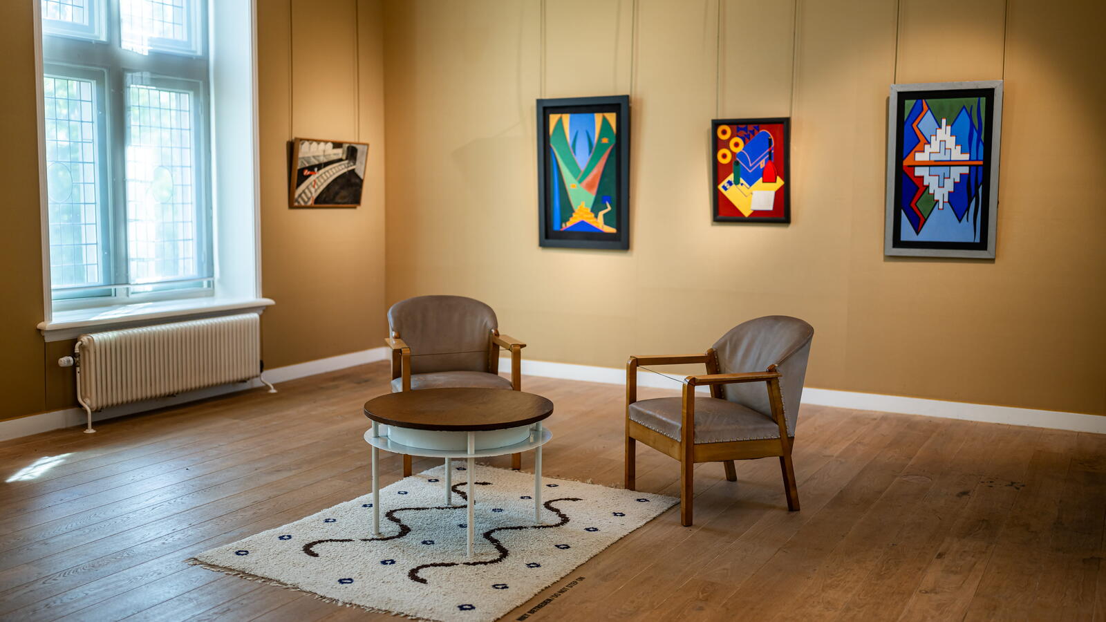 Een gele kamer met schilderijen aan de muren met abstracte thema's. Middenin de kamer staan twee stoelen, een ronde tafel en een tapijt.