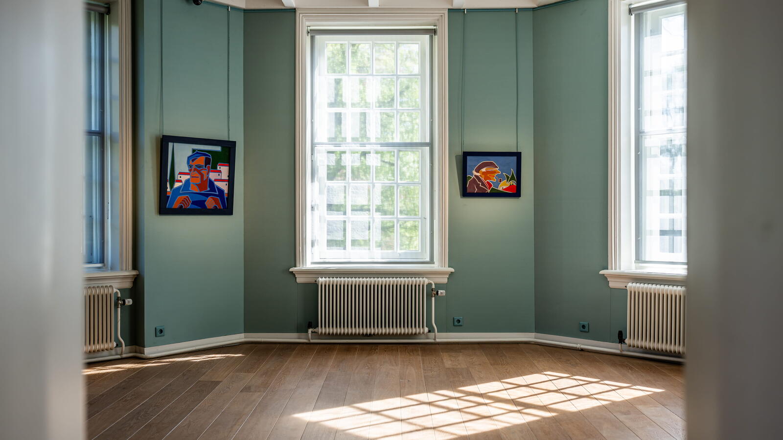 Een blauwe kamer met grote ramen en twee kleine schilderijen op de muur. Het licht valt op de houten vloer naar binnen.