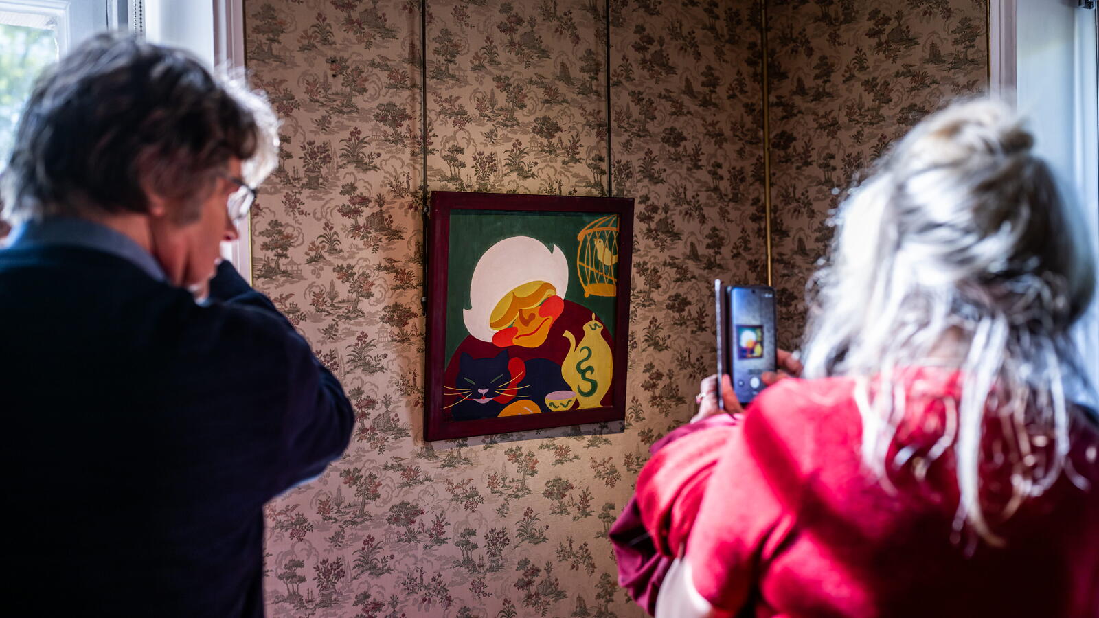 Een bezoeker in een roze jas neemt een foto van een kleurrijk schilderij van een oude vrouw met een zwarte kat op schoot.