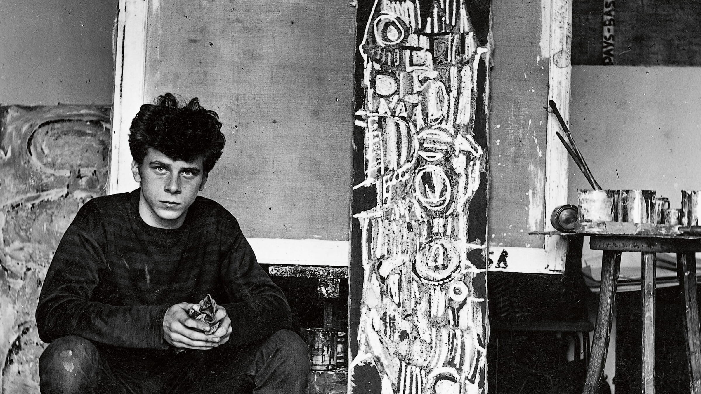 Een zwart-wit foto van een jonge Jan Cremer. Hij zit op een kruk omringd met verfspullen.