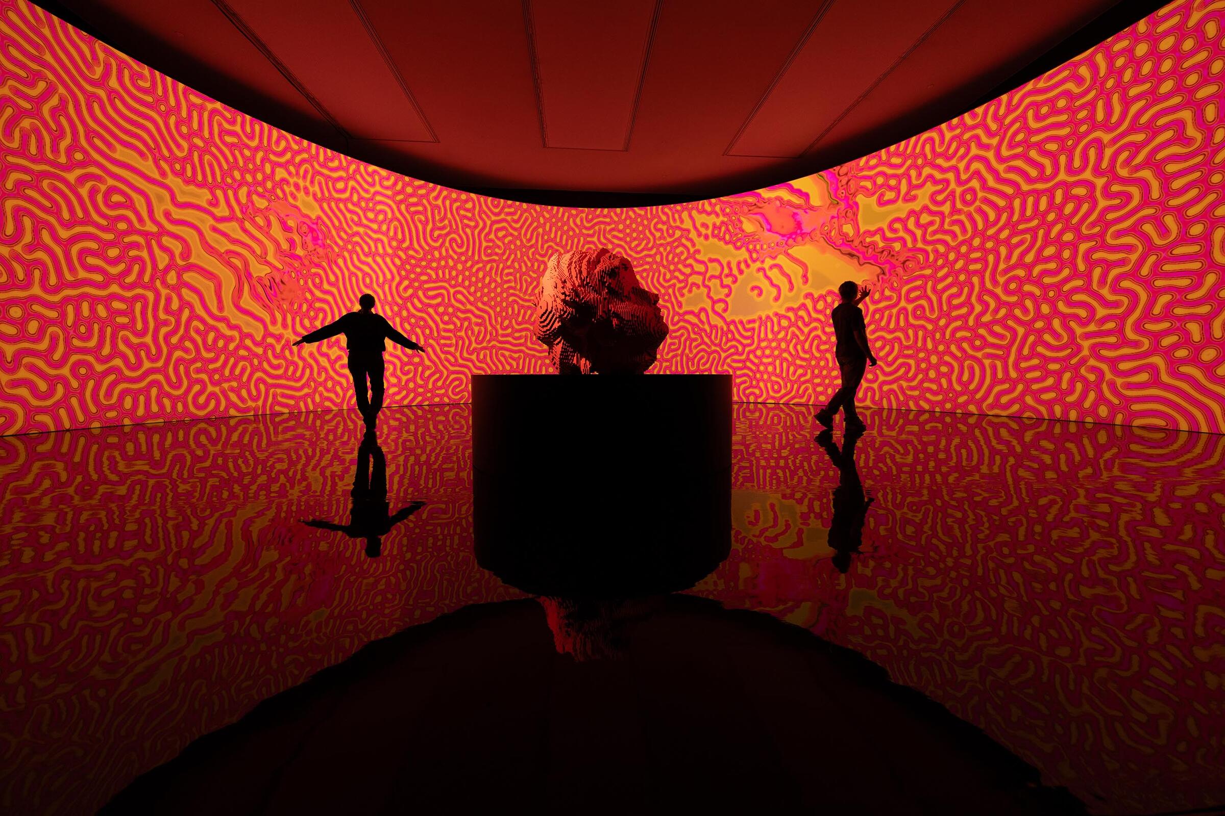 De muren zijn rood met allemaal kronkelende figuren in oranje en rood. Er zijn twee silhouetten van mensen die voor de schermen staan en zich uitstrekken ernaar. In het midden staat een standbeeld van een hoofd op een zwarte sokkel.