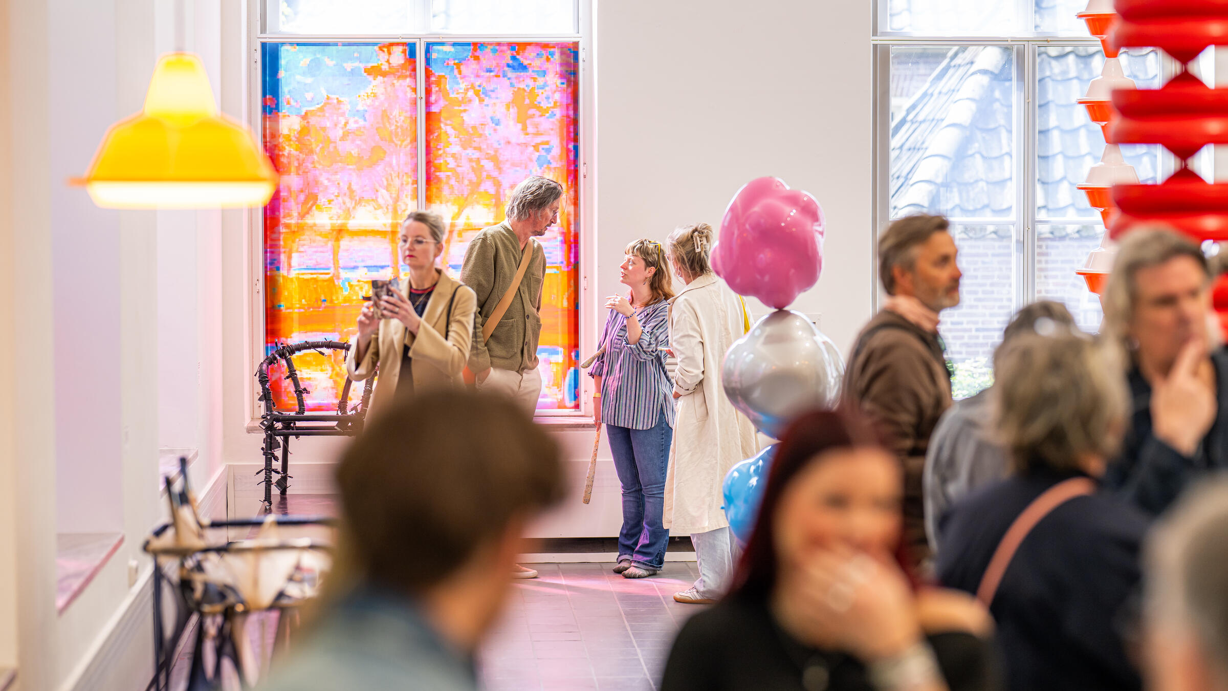 Bezoekers staan in een witte ruimte met kleurrijke kunstvoorwerpen. Een vrouw maakt een foto van een kunstwerk.