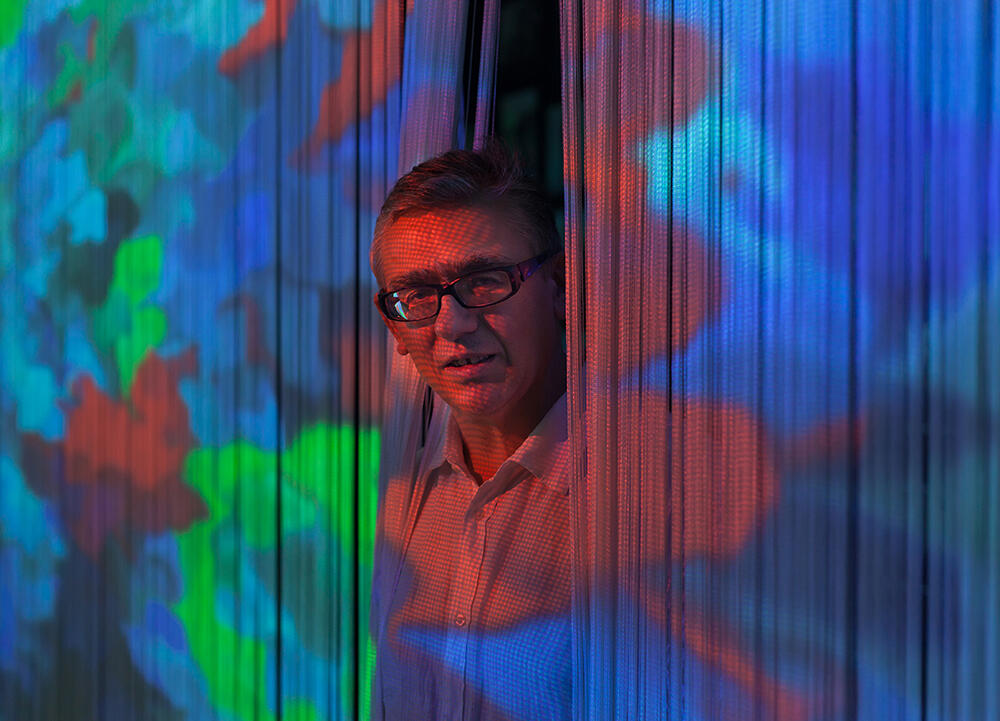 A portrait of Miguel Chevalier. He pokes his head through a curtain of thin strings on which a colourful digital artwork is projected.