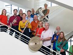 Muzikanten staan in een wit trappenhuis. Ze dragen allemaal verschillende kleuren shirts en houden hun instrumenten vast voor de foto. 