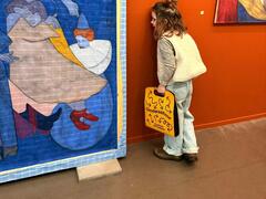 Een jong meisje staat met haar wang tegen de muur en kijkt naar de achterkant van een schilderij. In haar hand draagt ze een gele tas waar Omdwaaltour opstaat. 